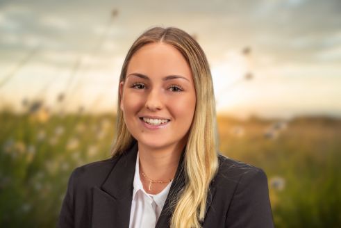 Freundlich lächelnde Frau, lange blonde Haare, schwarzes Jackett, Blumenwiese im Hintergrund.